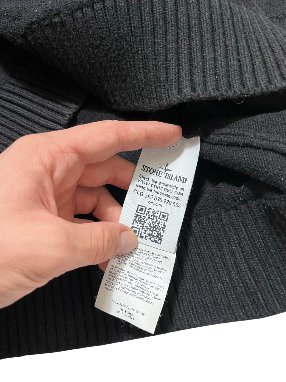 Stone Island címke fekete kötött pulóverben, kézben tartva, QR kóddal és márkajelzéssel.