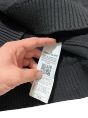 Stone Island címke fekete kötött pulóverben, kézben tartva, QR kóddal és márkajelzéssel.